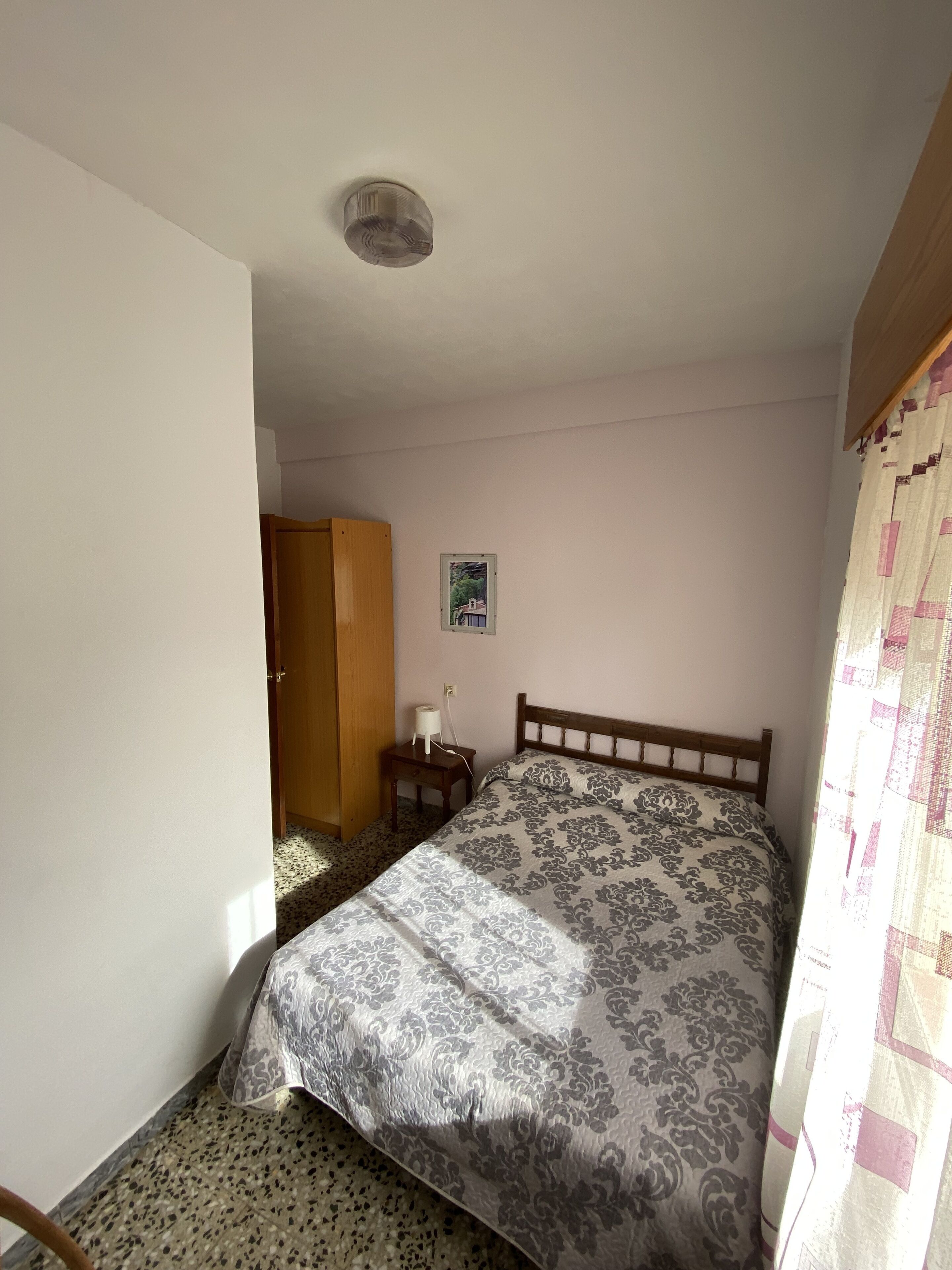 Un dormitorio con cama, mesita de noche, armario de madera, ventilador de techo y un cuadro pequeño en la pared.