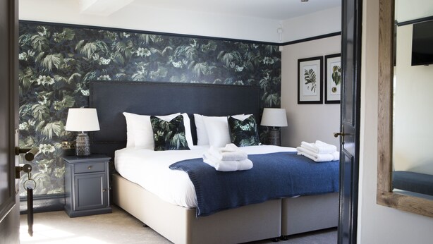 A bedroom with a large bed, two bedside tables with lamps, a floral wallpaper, and a wooden door. - Wynnstay Arms (Wrexham)