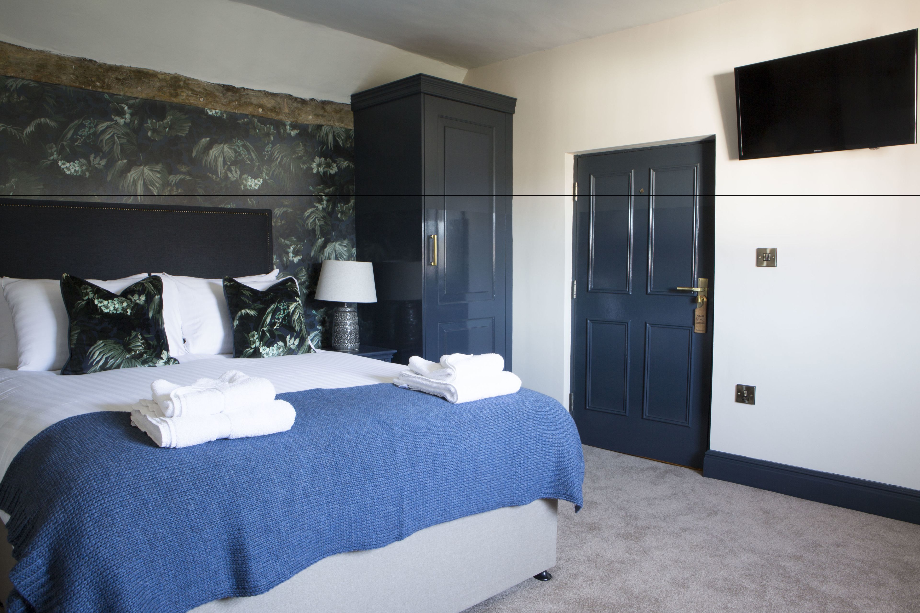 A neatly made bed with a blue blanket and white towels, a dark blue wardrobe, a lamp, and a television on the wall.