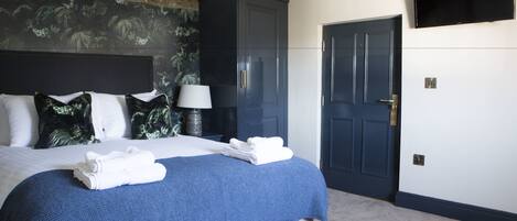 A neatly made bed with a blue blanket and white towels, a dark blue wardrobe, a lamp, and a television on the wall.