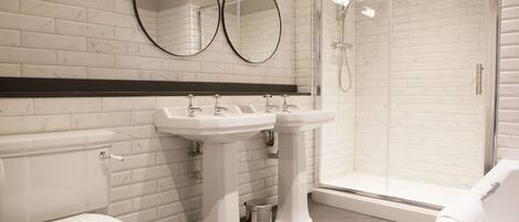 Un baño moderno con zócalos de mármol blanco, dos lavamanos, inodoro y ducha con mampara de vidrio.