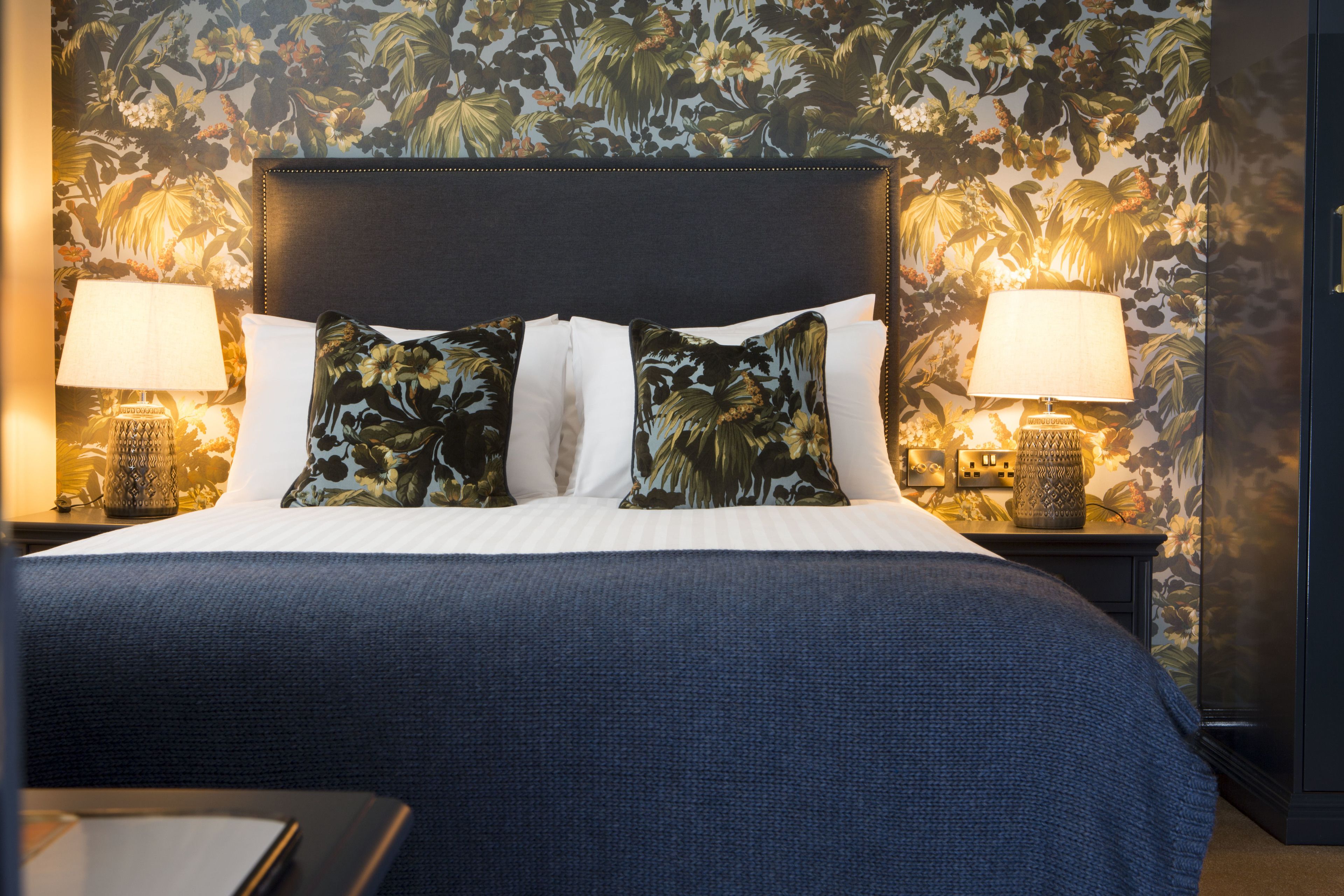 Una cama bien hecha con almohadas de estampado floral y un cabecero oscuro, flanqueada por dos lámparas de noche sobre mesitas a juego.