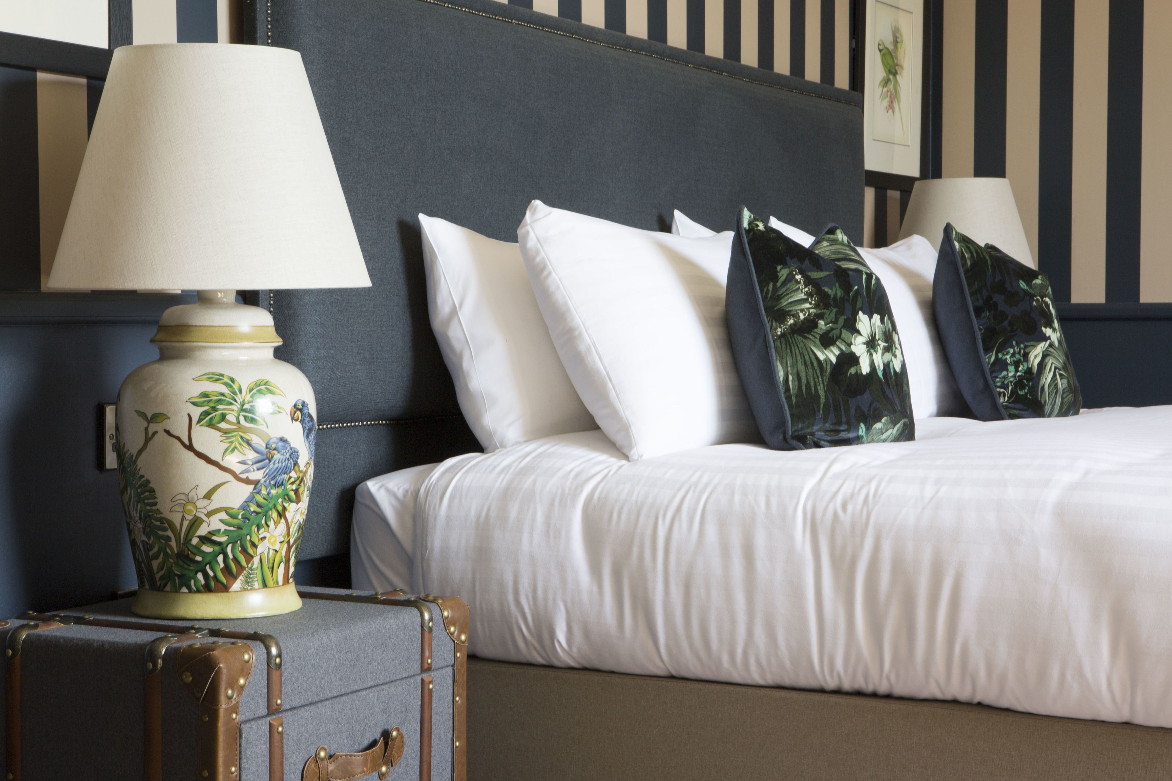 Een net opgemaakt bed met witte lakens en decoratieve kussens, een lamp met bloemenmotief, en een vintage koffer op de vloer.