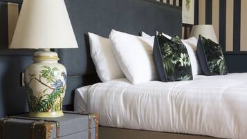 Een net opgemaakt bed met witte lakens en decoratieve kussens, een lamp met bloemenmotief, en een vintage koffer op de vloer.