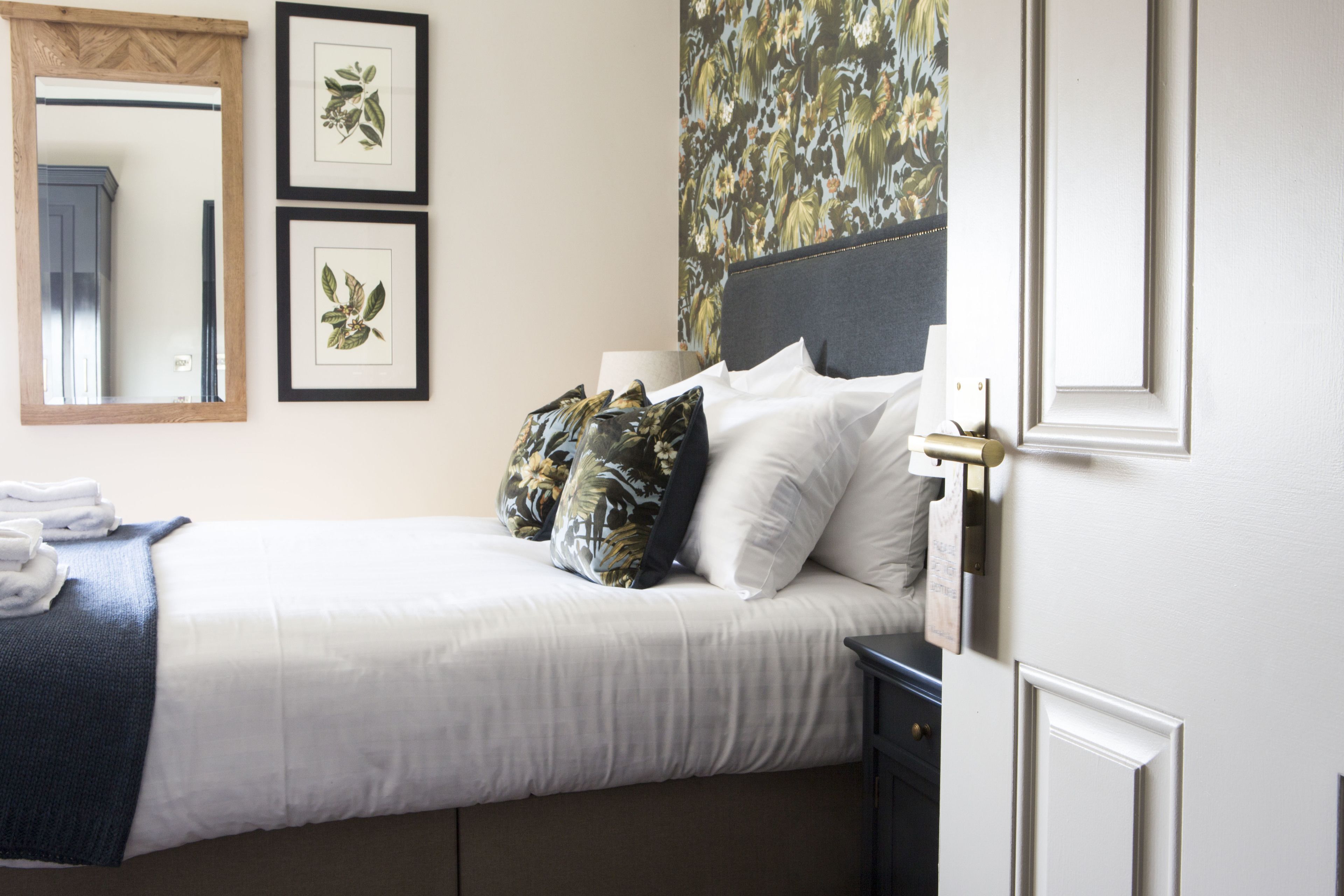 A neatly made bed with white linens, a dark headboard, and two framed botanical prints on the wall.