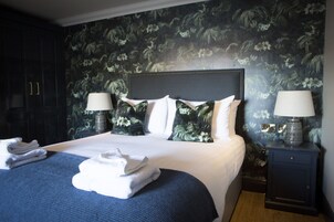 Una cama bien hecha con un cabecero oscuro, dos lámparas de noche y un papel tapiz con motivos florales.