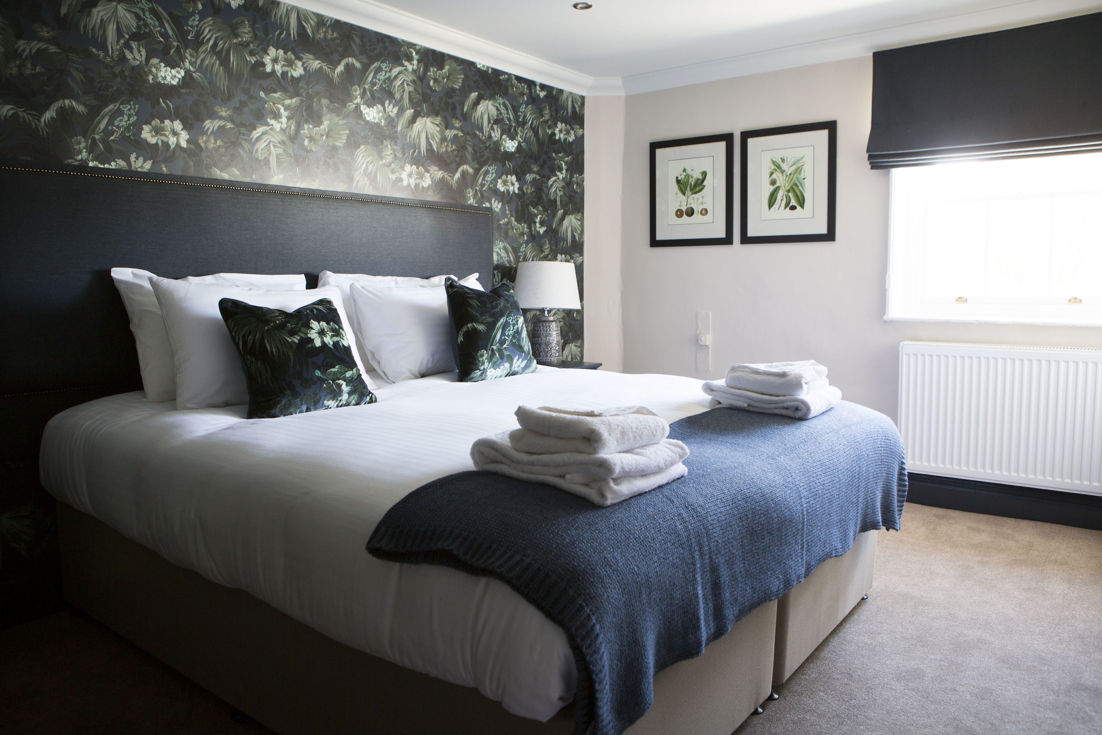 A neatly made bed with white linens and a blue throw blanket, a bedside lamp, and two framed botanical prints on the wall.