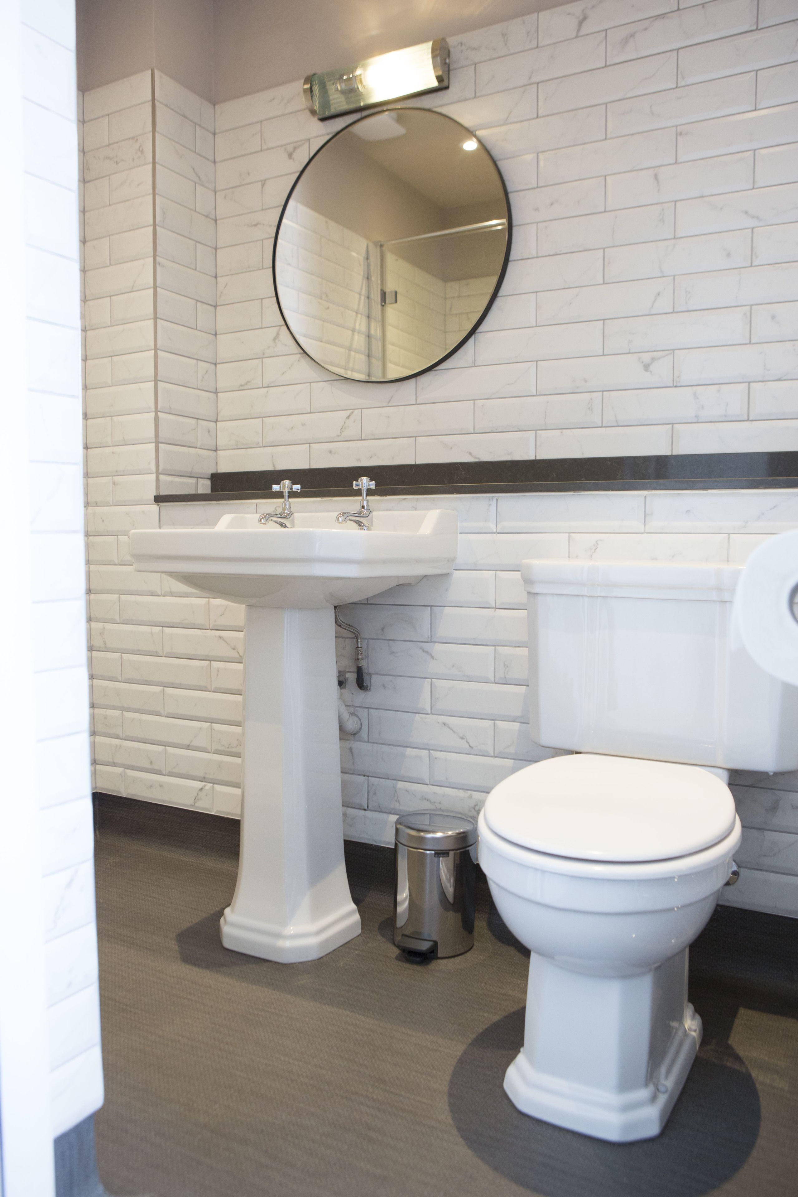 Une salle de bain avec un lavabo sur colonne blanc, un miroir rond, des toilettes et une poubelle.