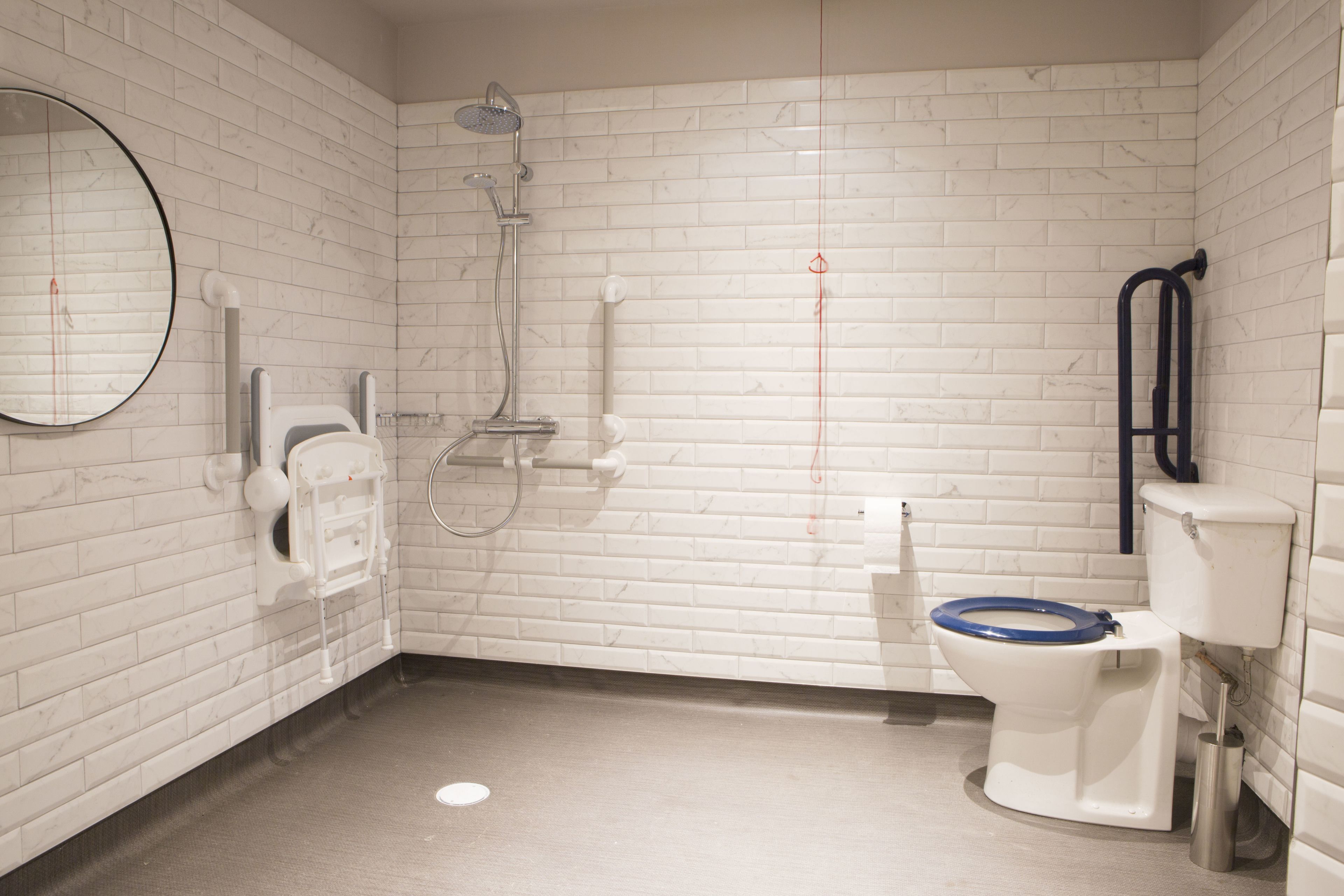 A bathroom with white brick walls, a white toilet with a blue seat, a showerhead, a hand dryer, and a mirror.