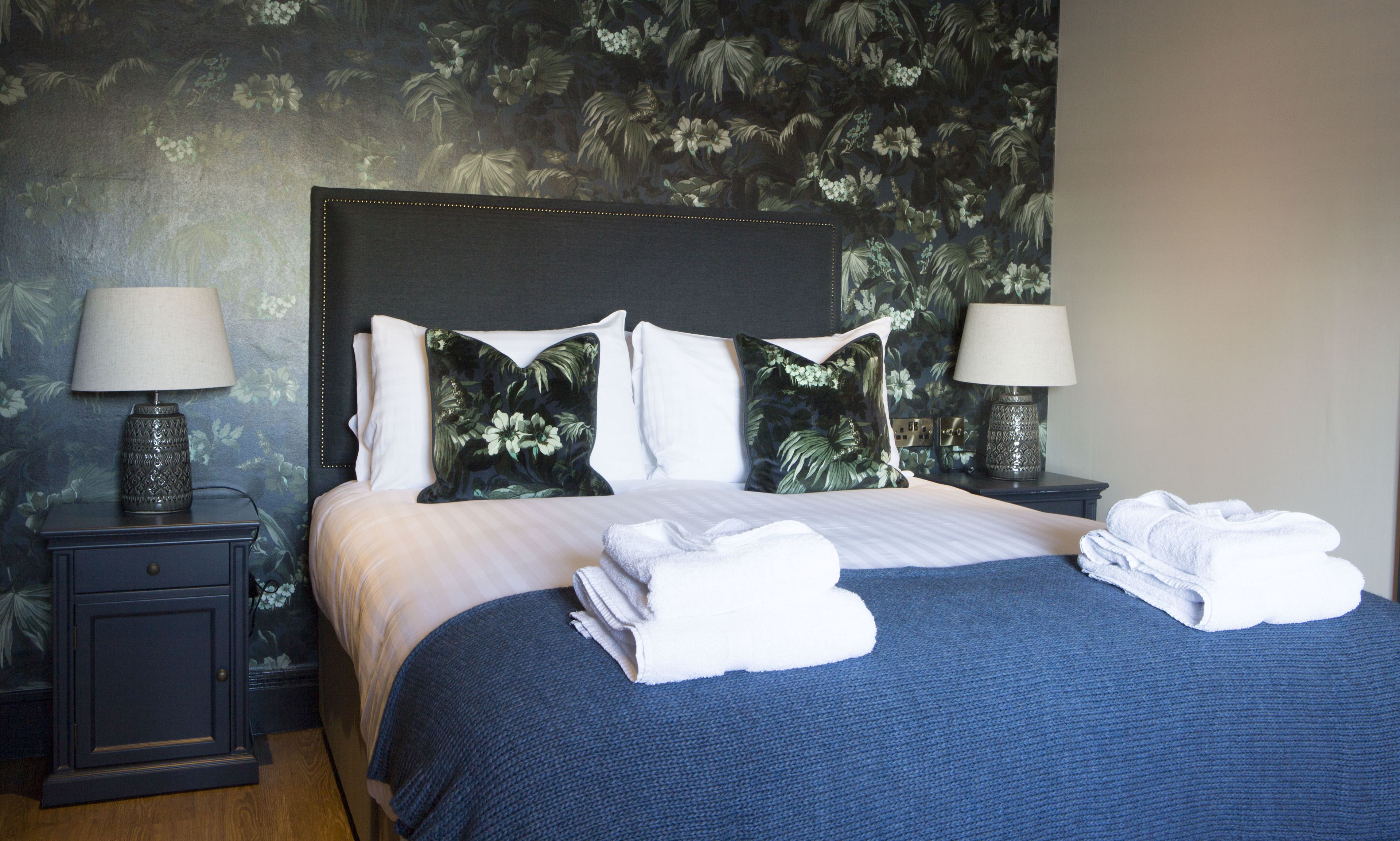 A neatly made bed with a blue blanket and white pillows, two bedside tables with lamps, and a floral wallpaper.