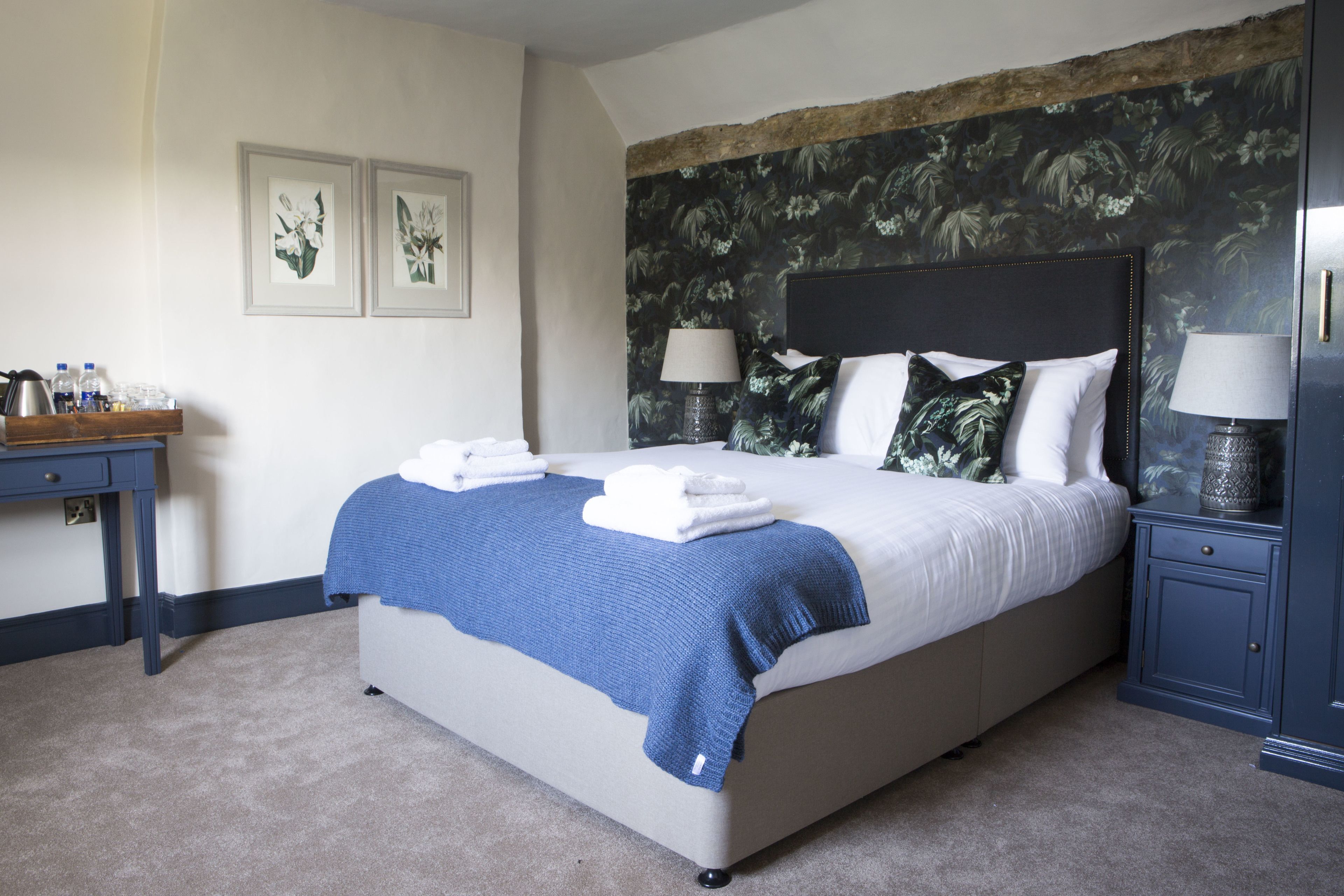 A neatly made bed with a blue blanket, a bedside table with a lamp, and a wall with floral wallpaper.