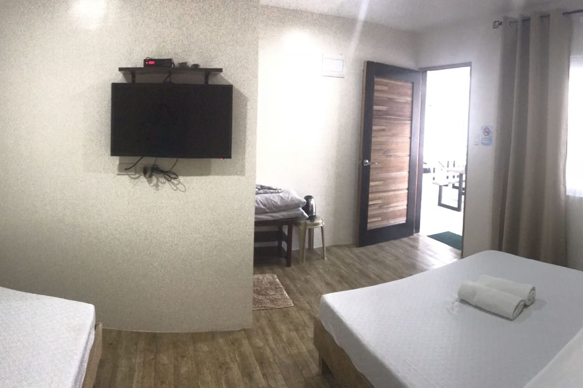 A hotel room with two beds, a TV, a wall-mounted heater, and a view of the outside.