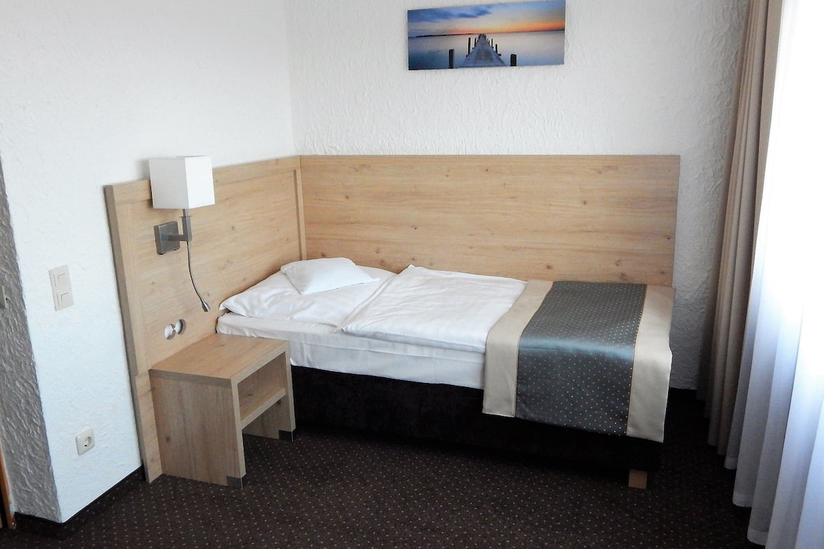Ein Hotelzimmer mit massivem Holzkopfteil, einem Einzelbett, einem Nachttisch, einer Wandleuchte und einem Gemälde, das einen Sonnenuntergang über einem Steg zeigt.