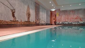 Piscina interior com piso ladrilhado, parede de pedra com mural de montanhas e teto com iluminação embutida.