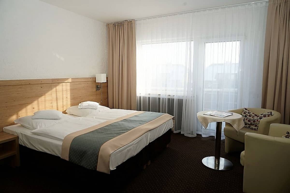 Ein Hotelzimmer mit Bett, kleinem Tisch, Stuhl und einem Fenster mit Vorhängen.
