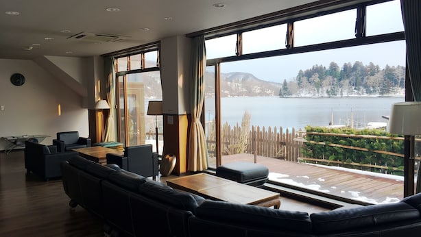 A modern living room with a large window overlooking a lake, furnished with a sofa, coffee table, and lamps. - Nojiri Lake Resort (Shinano)