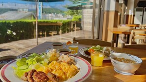 A table with breakfast items including eggs, bacon, and vegetables, with a view of a garden and hills in the background. - Nojiri Lake Resort (Shinano)