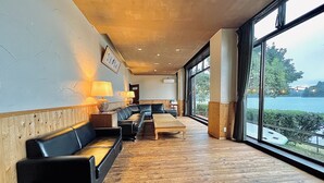 A spacious room with a wooden floor, a black leather sofa, a wooden table, and large windows offering a view of a body of water. - Nojiri Lake Resort (Shinano)