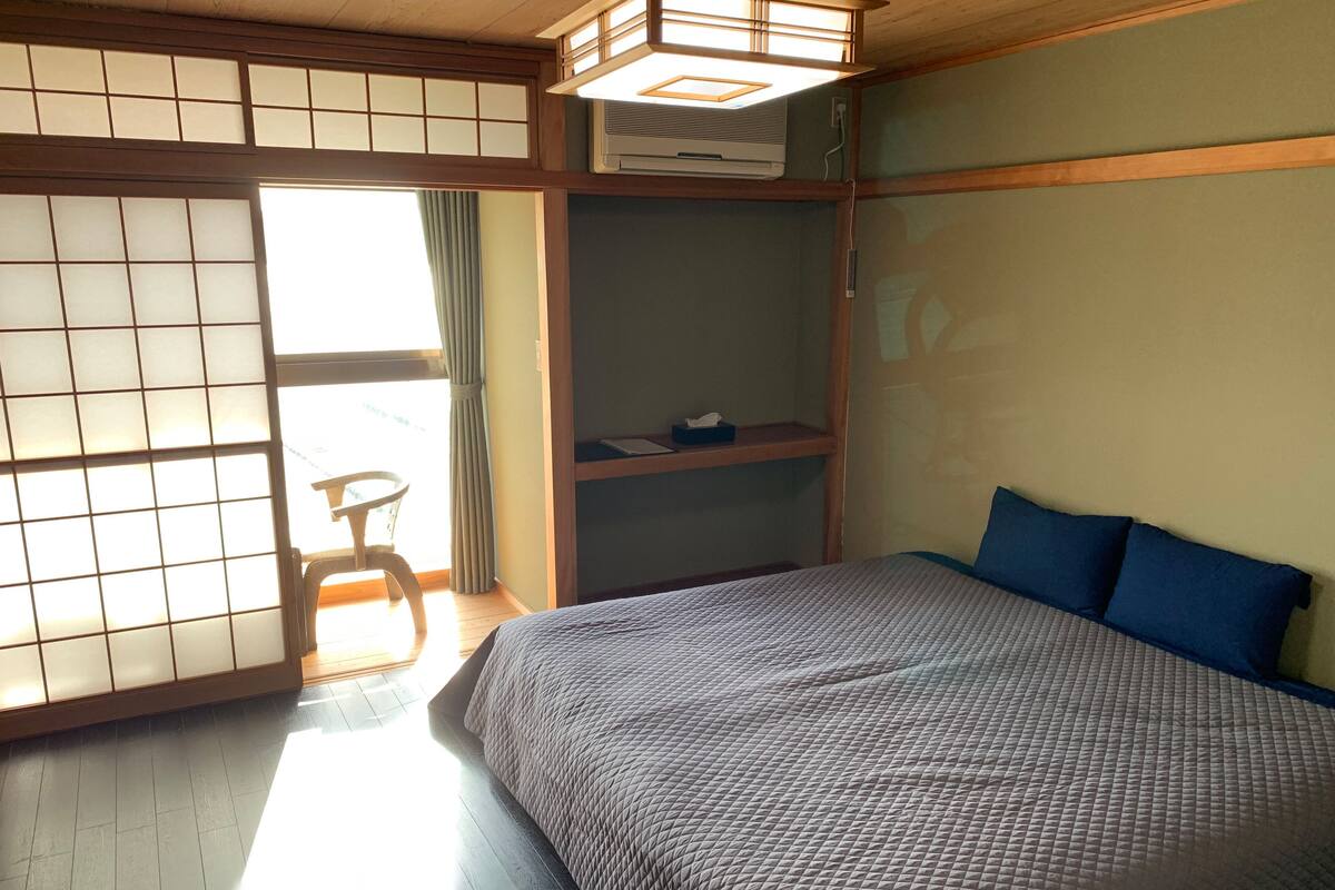 A traditional Japanese-style room with tatami flooring, a low bed with blue pillows, a futon, a sliding shoji screen door, and a small table with a chair.