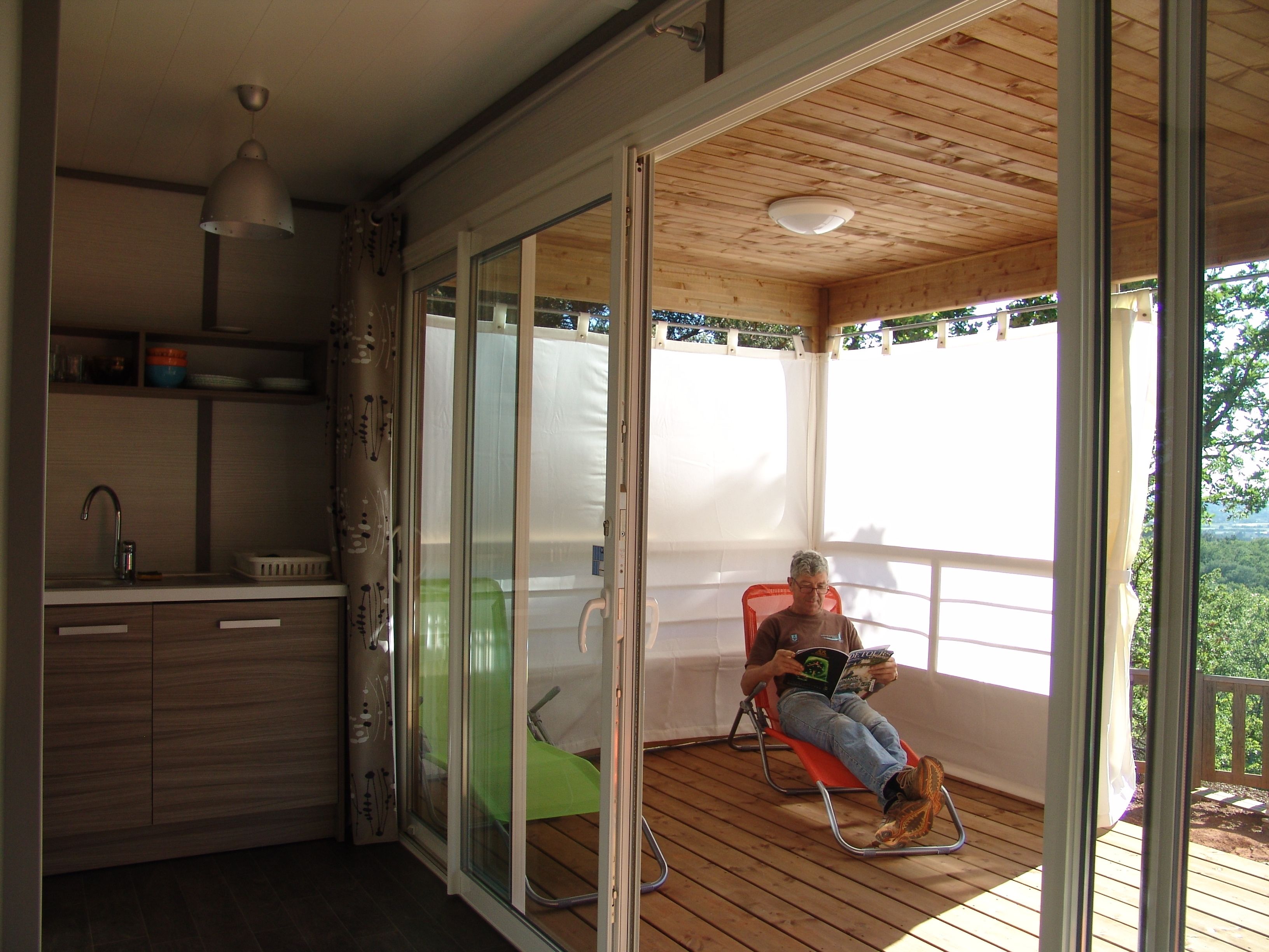 Una habitación con techo de madera, una puerta corrediza de vidrio que da a un espacio exterior, una pequeña cocina americana y una persona sentada en una silla leyendo un libro.