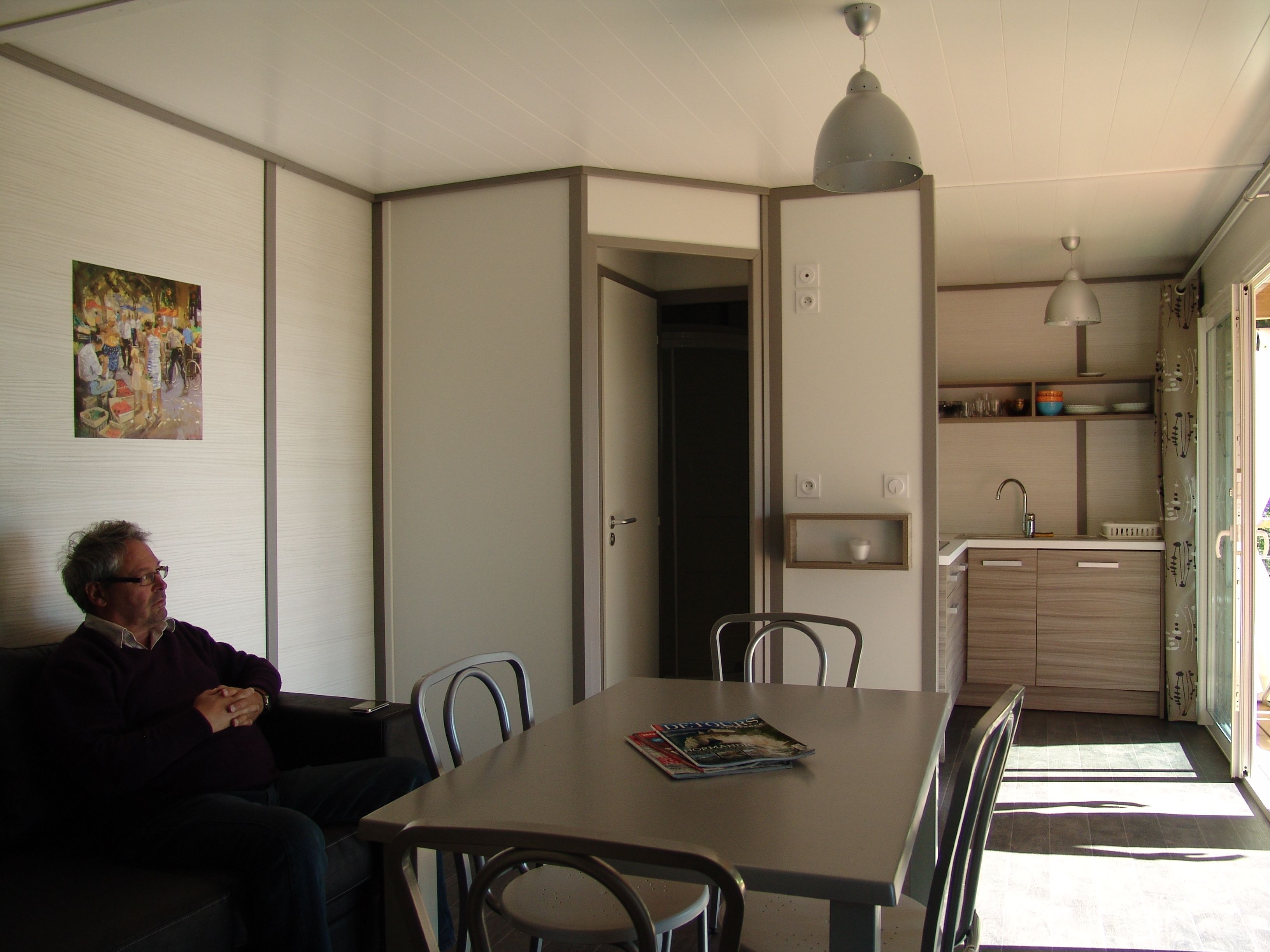 A living room with a sofa, a dining table, and chairs. There is a painting on the wall and a kitchen area visible through an open door.