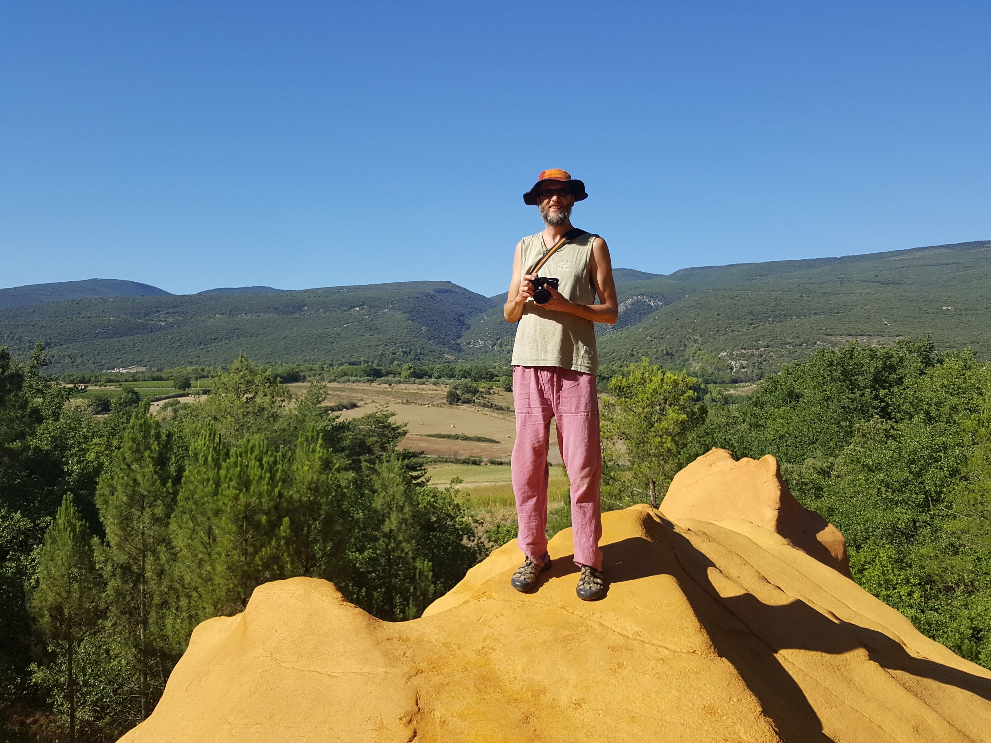 A person standing on a sandy mound with a camera, overlooking a landscape with hills and trees.
