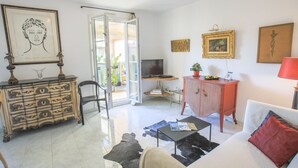 A living room with a white sofa, a coffee table, a red cabinet, and a television. There is a framed picture on the wall and a chandelier. - COLLINE DU CHATEAU AP4077 by RIVIERA HOLIDAY HOMES (Niza)