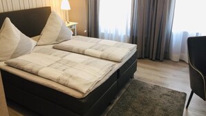 A neatly made bed with a checkered bedspread and pillows, a bedside lamp, and a chair in a room with a wooden floor and curtains. - Gästehaus Albers (Cochem)