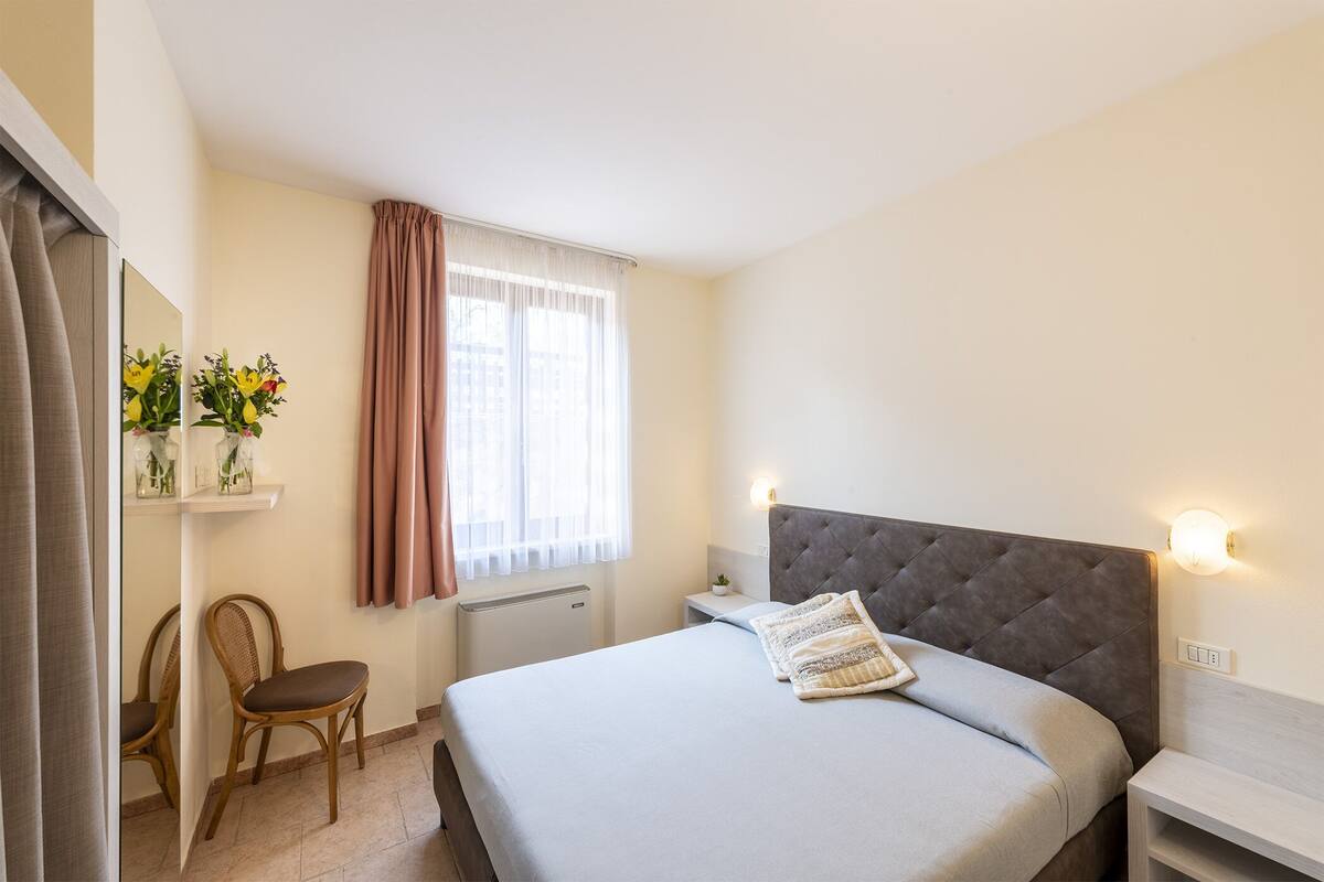 A bedroom with a bed, a chair, a nightstand, a wall-mounted light, and a window with curtains.