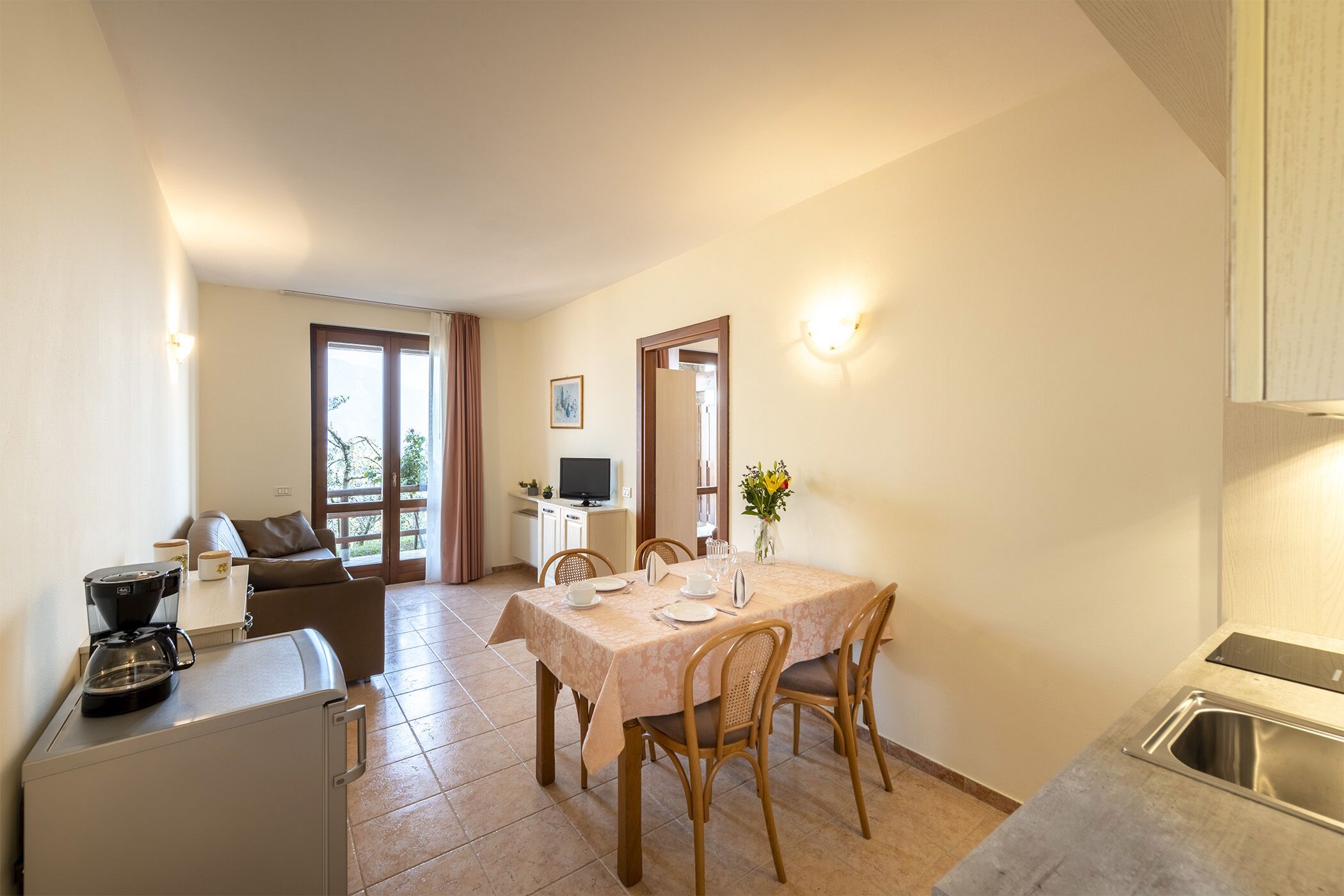 A compact kitchen with a dining area, a small refrigerator, a coffee maker, and a TV in the background.