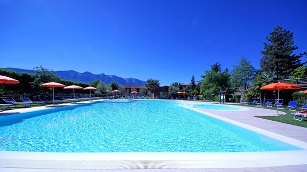 A large outdoor swimming pool with lounge chairs and umbrellas, surrounded by trees and mountains in the background. - Residence Pineta Campi (Tremosine)