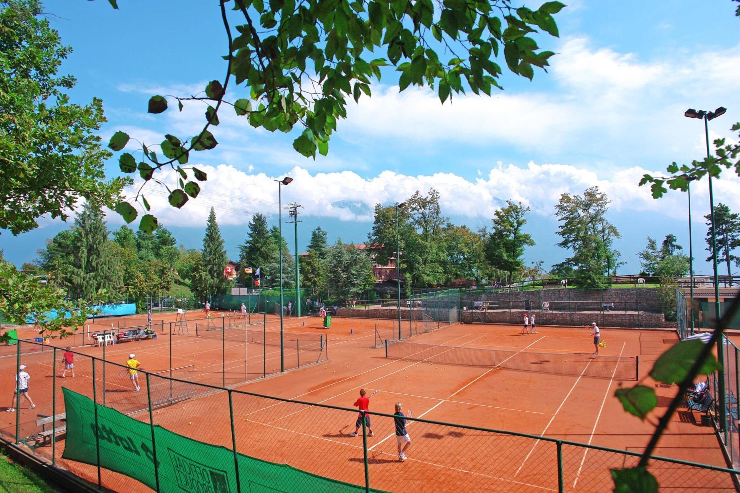 Cancha de tenis con varios jugadores, rodeada de árboles y un cielo despejado.