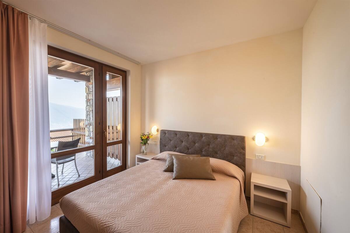 A hotel room with a large bed, a nightstand, a wall-mounted light, and a view of the outdoors through a sliding door.