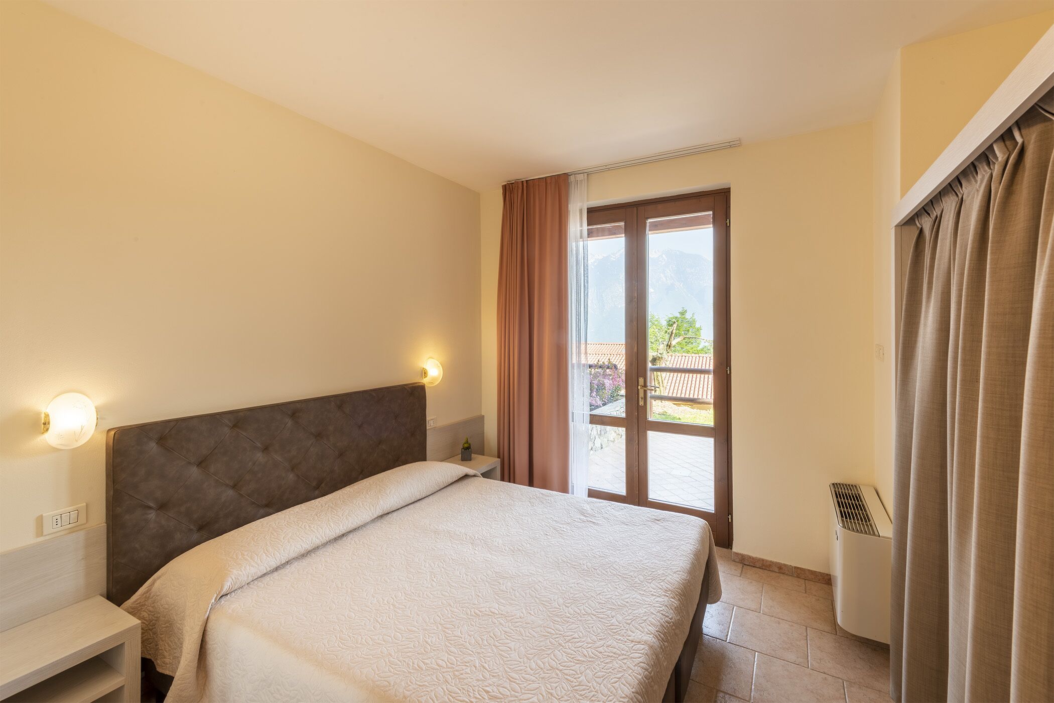 A hotel room with a bed, a nightstand, a wall-mounted light, a sliding glass door to an outdoor area, and an air conditioning unit.