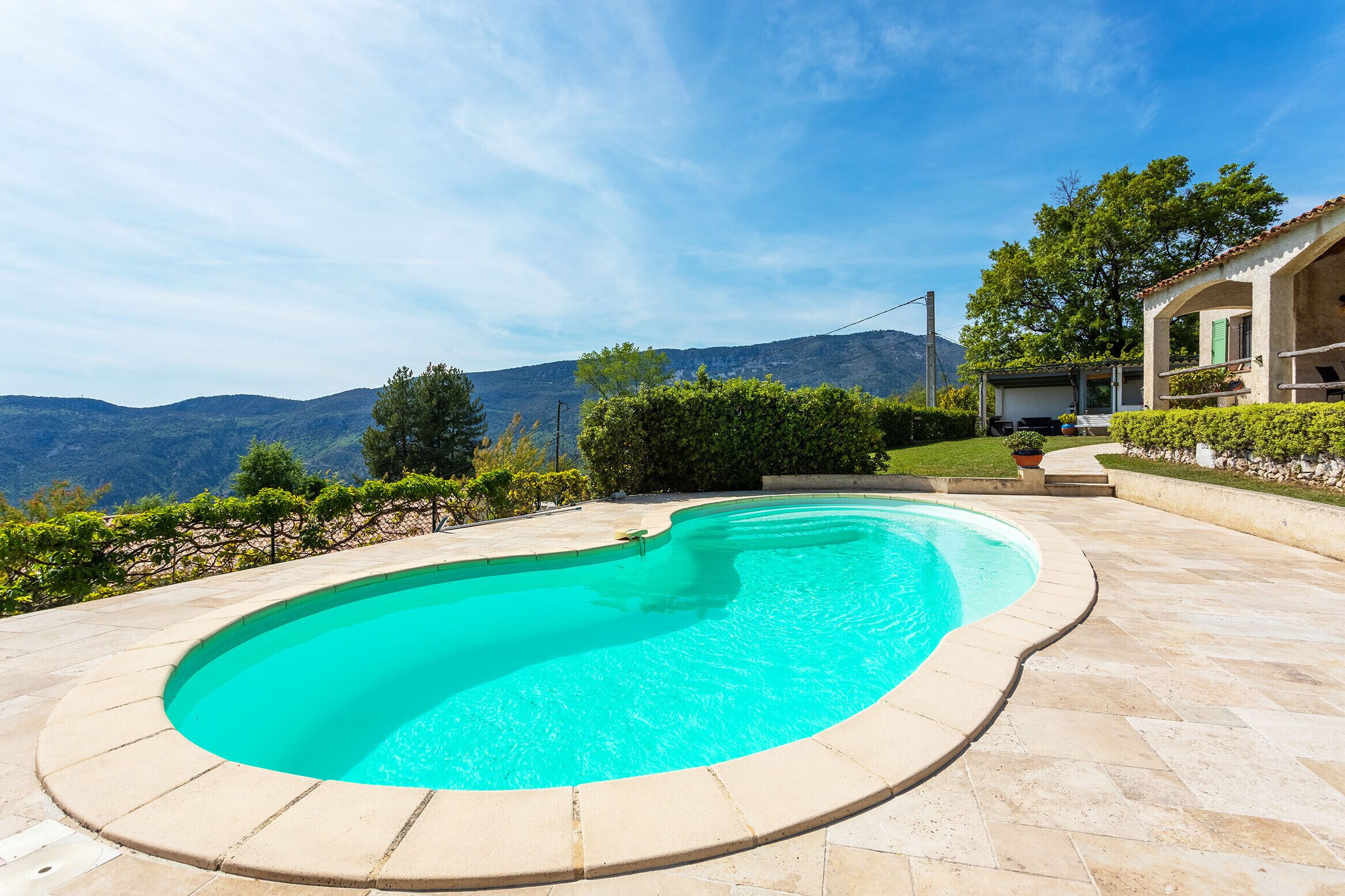 A swimming pool with a curved edge, surrounded by a stone patio, and a house with a tiled roof in the background.