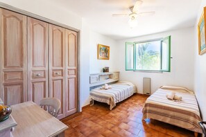 Ein Schlafzimmer mit zwei Betten, einem Holzschrank, einem Schreibtisch mit Stuhl, einem Heizkörper und einem Fenster mit Blick ins Grüne.