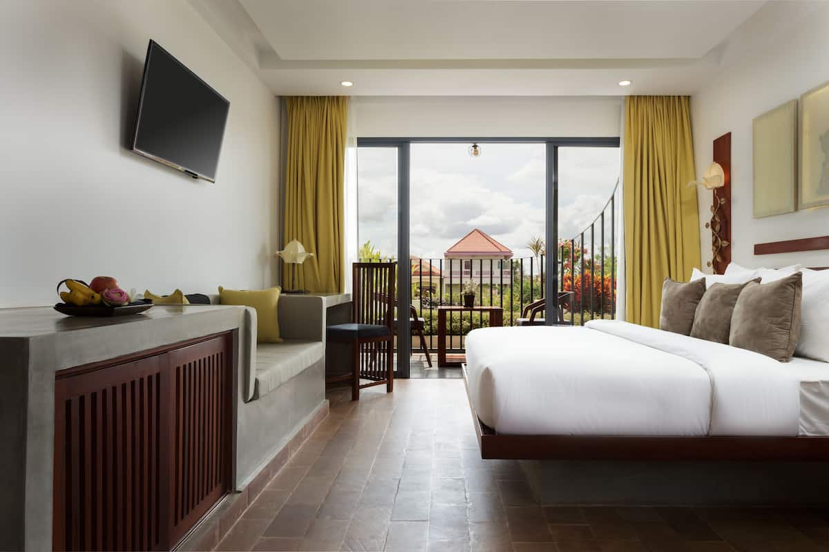 A hotel room with a bed, a desk with a flat-screen TV, a balcony with a view of the outside, and a dining area with chairs and a table.