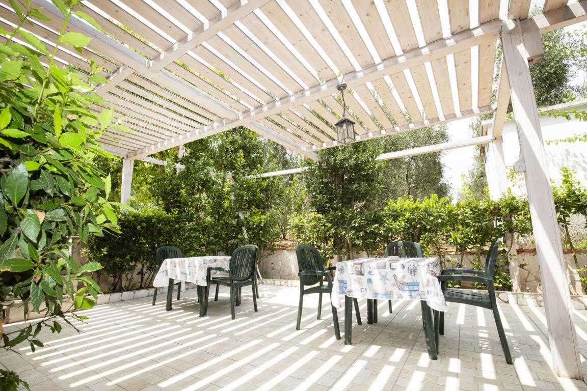 An outdoor patio with wooden pergola, green chairs, and tables covered with white tablecloths.