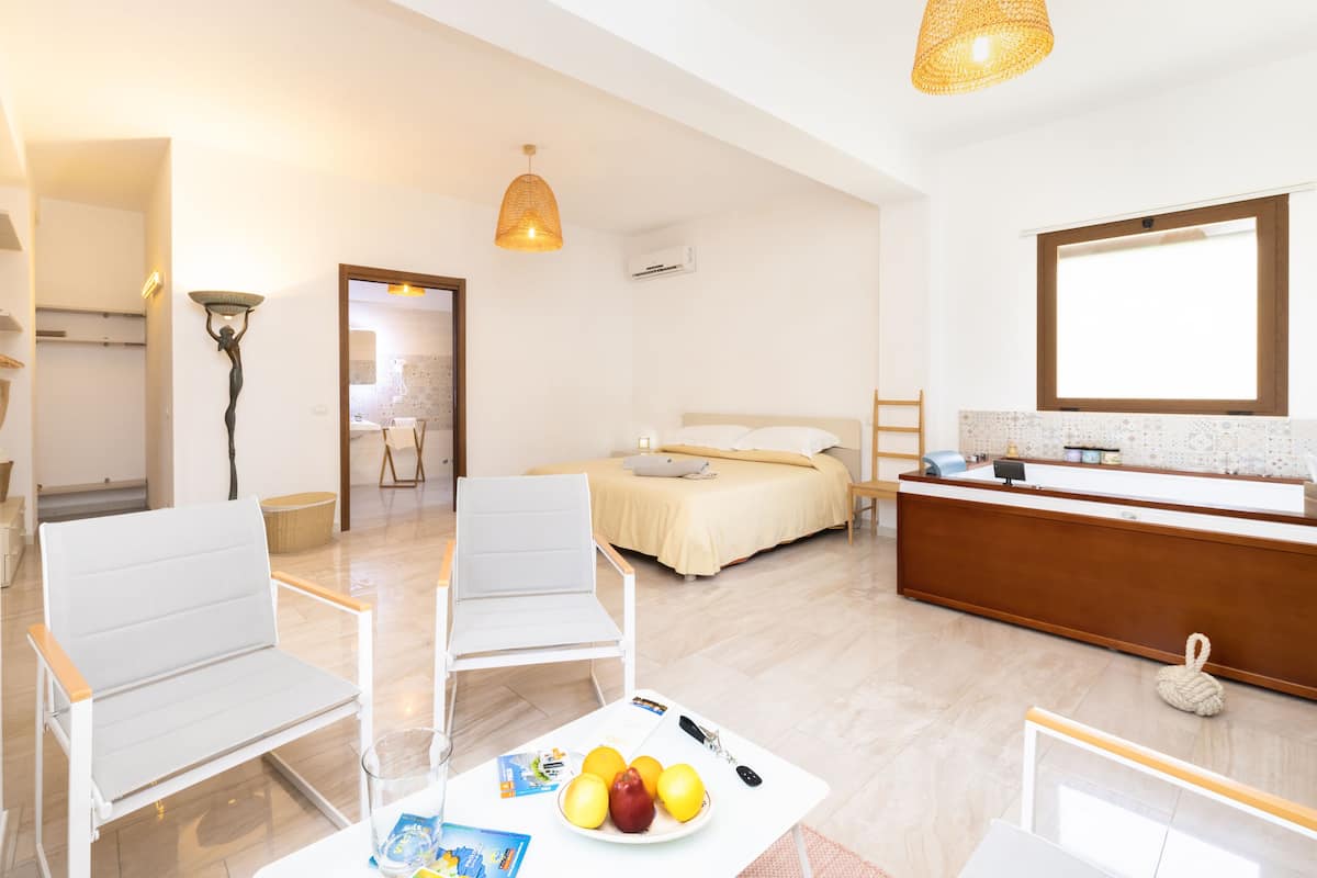 A modern hotel room with a bed, two white chairs, a dining table with fruit and brochures, a desk with a mirror, and a hanging light fixture.
