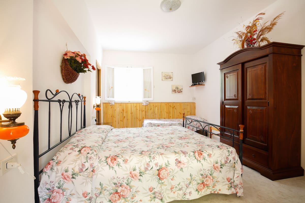 A bedroom with a wooden headboard, a floral bedspread, a wooden wardrobe, and a bedside table with a lamp.