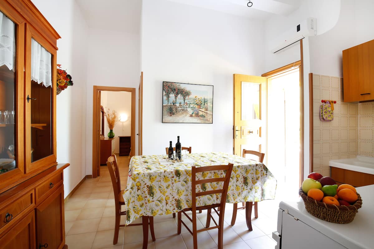 A dining area with a table set for two, a cabinet with glass doors, a painting on the wall, and a kitchen area with fruit on the counter.