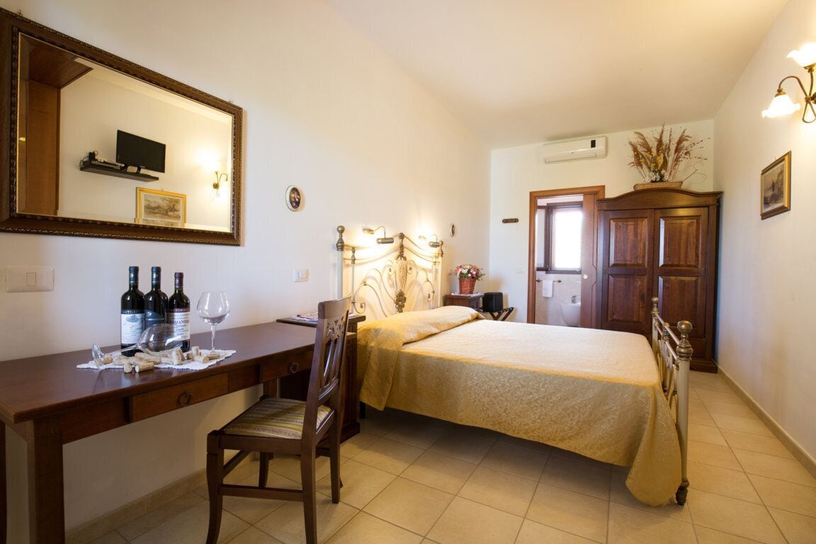 A bedroom with a bed, a desk with wine bottles, a chair, a dresser, and a mirror.