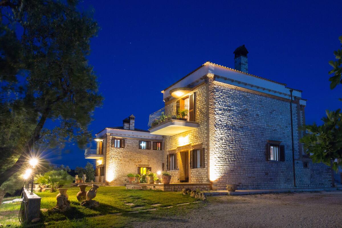 A stone building with a balcony and multiple windows, surrounded by trees and a well-maintained lawn.