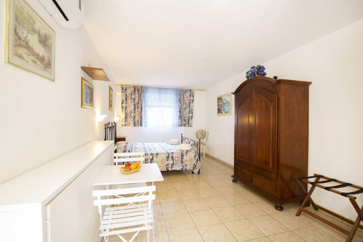 A small, bright room with a bed, a wooden wardrobe, a table with fruit, and a window with floral curtains.