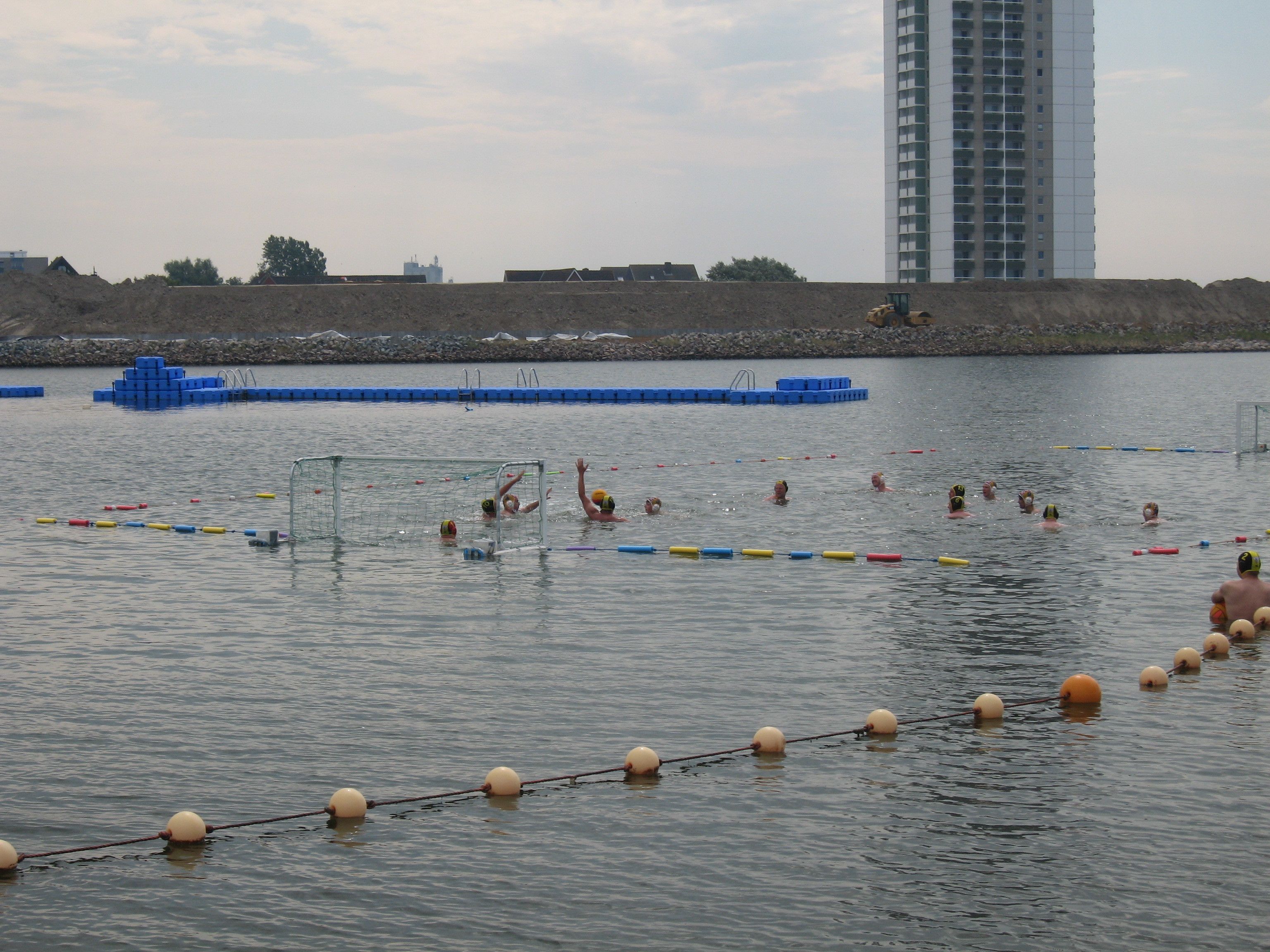 A water polo game in a large outdoor pool with a goal, surrounded by water polo balls and buoys.