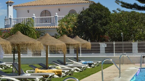 Zona de piscina con sillones reclinables y sombrillas de paja, un edificio blanco con balcón y piscina cercada.