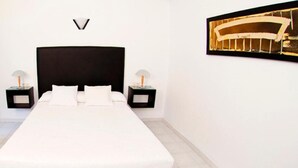 Un dormitorio con una cama grande, dos mesitas de noche con lámparas, una puerta y un cuadro enmarcado en la pared.