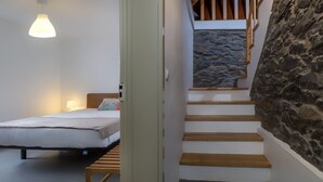 Un dormitorio con cama, lámpara de noche y un banco de madera. Se ve una escalera de madera con escalones de piedra y una pared de piedra.
