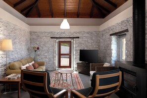 A cozy living room with a stone wall, wooden ceiling beams, a sofa, a TV, a fireplace, and a dining area with chairs and a table.