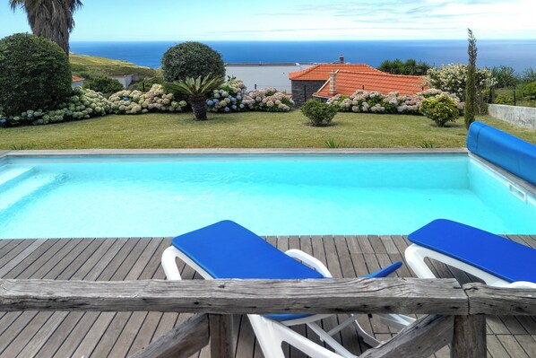 A swimming pool with two lounge chairs, surrounded by a wooden deck and a view of the sea.